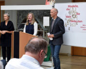 Drei Personen stehen während einer Präsentation vor einem Plakat der Hochschule Coburg. Das Plakat zeigt einen deutschen Text und ein Motiv mit Figuren und Bäumen. Im Vordergrund sitzen Zuhörer und hören aufmerksam zu.