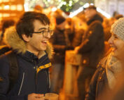 Zwei Menschen in warmer Kleidung genießen eine lebhafte, illuminierte Veranstaltung an der Hochschule Coburg, lächeln und halten warme Getränke in den Händen. Der Hintergrund ist mit festlichen Lichtern und einer Menge verschwommenen Figuren gefüllt, was eine fröhliche und gemütliche Atmosphäre schafft.