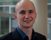 Eine lächelnde Person in einem dunklen Blazer und einem blauen Hemd steht in einem Innenbereich der Hochschule Coburg vor großen Fenstern und einer Säule. Im Hintergrund ist ein modernes Interieur mit verschwommenen architektonischen Details zu sehen.