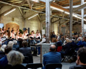 Ein Chor der Hochschule Coburg tritt in einem rustikalen Open-Air-Saal mit freiliegenden Balken und Backsteinwänden auf. Der Dirigent steht vor dem Chor, während das sitzende Publikum die Aufführung aufmerksam verfolgt und die Stuhlreihen füllt.