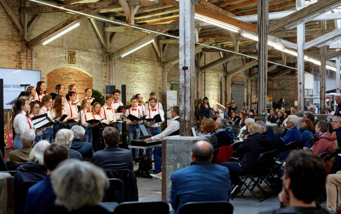 Ein Chor der Hochschule Coburg tritt in einem rustikalen Open-Air-Saal mit freiliegenden Balken und Backsteinwänden auf. Der Dirigent steht vor dem Chor, während das sitzende Publikum die Aufführung aufmerksam verfolgt und die Stuhlreihen füllt.