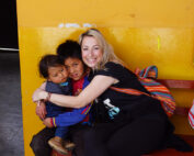 Eine lächelnde Frau mit einem bunten Rucksack umarmt zwei Kinder auf einer roten Bank vor einer leuchtend gelben Wand der Hochschule Coburg. Die Kinder sehen zufrieden aus und sind leger gekleidet, was der Szene Wärme und Kameradschaft verleiht.