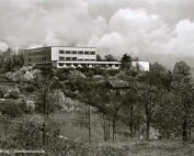 Ein Schwarzweißfoto eines großen rechteckigen Gebäudes mit der Aufschrift „Coburg – Staatsbauschule“ auf einer Hügelspitze, umgeben von Bäumen und Sträuchern unter einem bewölkten Himmel, das an die historischen Wurzeln der Hochschule Coburg erinnert.