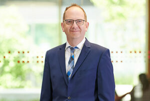 A person wearing glasses and a blue suit with a patterned tie stands smiling in front of the Hochschule Coburg, with its distinct architecture softly blurred in the background.
