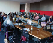 Eine Gruppe von Mitarbeitern der Hochschule Coburg sitzt in einem modernen Büro um einen großen U-förmigen Konferenztisch. Jeder hat einen Laptop, auf Bildschirmen laufen Präsentationsfolien. Der Raum ist in Rot und Grau gehalten, im hinteren Bereich gibt es Erfrischungen.