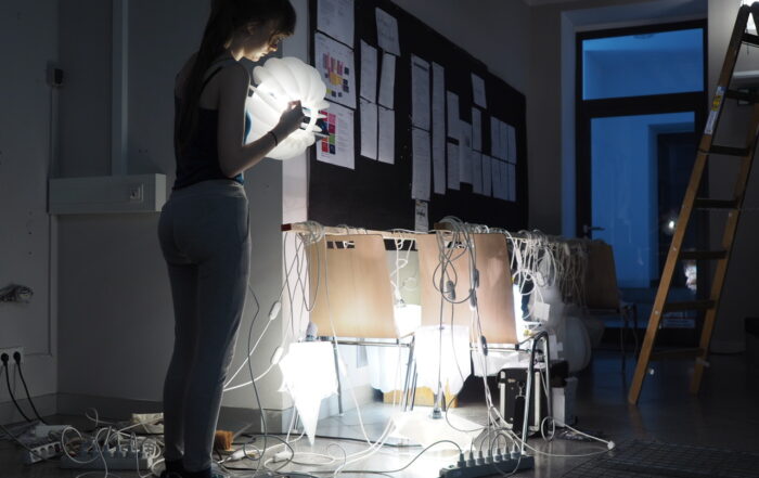 In einem schwach beleuchteten Raum der Hochschule Coburg steht eine Person und hält eine runde Lampe in der Hand. Mehrere Stühle sind mit verhedderten weißen Kabeln und Glühbirnen übersät. Eine große schwarze Pinnwand mit Papieren ziert die Wand, und daneben steht eine hohe Leiter, bereit für ihre nächste Aufgabe.