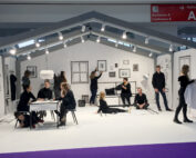 Eine Gruppe schwarz gekleideter Menschen beschäftigt sich in einem weißen Raum der Hochschule Coburg, der wie eine 2D-Zeichnung aussieht, mit verschiedenen Aktivitäten. Einige sitzen, stehen und interagieren mit dem minimalistisch illustrierten Dekor.