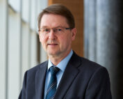 Ein Mann mit kurzen braunen Haaren und Brille, der einen dunklen Anzug, ein hellblaues Hemd und eine blau gestreifte Krawatte trägt, steht in einem Gebäude der Hochschule Coburg. Er steht vor einem unscharfen Hintergrund mit großen Fenstern und einer Säule.