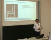 Eine Frau steht vor einem Hörsaal der Hochschule Coburg und hält für Rohde & Schwarz einen Vortrag zum Thema „Change Management“. Auf einer großen Leinwand werden zentrale Themen wie Teamentwicklung und Moderation präsentiert. Im Vordergrund komplettieren eine Tafel und ein Laptop die dynamische Lernumgebung.