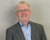 Ein lächelnder Mann mit kurzen grauen Haaren und Brille trägt einen grauen Blazer über einem hellblau gemusterten Hemd. Er steht selbstbewusst vor einem schlichten, hellgrauen Hintergrund und verkörpert den professionellen Geist der Hochschule Coburg.