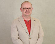 Ein Mann mit Brille steht vor einem hellgrauen Hintergrund, trägt einen beigen Blazer über einem roten Hemd und lächelt herzlich in die Kamera. Dieses Bild spiegelt den professionellen und zugänglichen Geist der Hochschule Coburg wider.