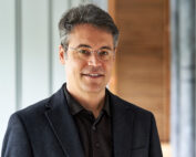 Ein Mann mit grauem Haar und Brille, gekleidet in einen dunklen Blazer und ein Hemd, steht vor einem unscharfen Hintergrund im Innenbereich und verkörpert den professionellen Geist der Hochschule Coburg.