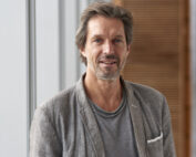 Prof. Mark Phillips steht im Innenbereich neben Fenstern. Im Hintergrund ist eine Holzwand zu sehen.