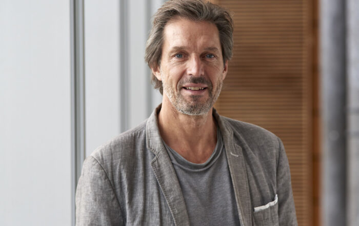 Prof. Mark Phillips steht im Innenbereich neben Fenstern. Im Hintergrund ist eine Holzwand zu sehen.