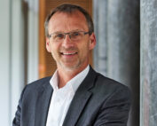 Ein Mann in Anzug und weißem Hemd steht lächelnd in einem modernen Innenraum der Hochschule Coburg. Er hat kurze Haare, trägt eine Brille und hat die Arme verschränkt. Im Hintergrund sind Säulen und verschwommene Elemente zu sehen, die an eine Büroumgebung erinnern.