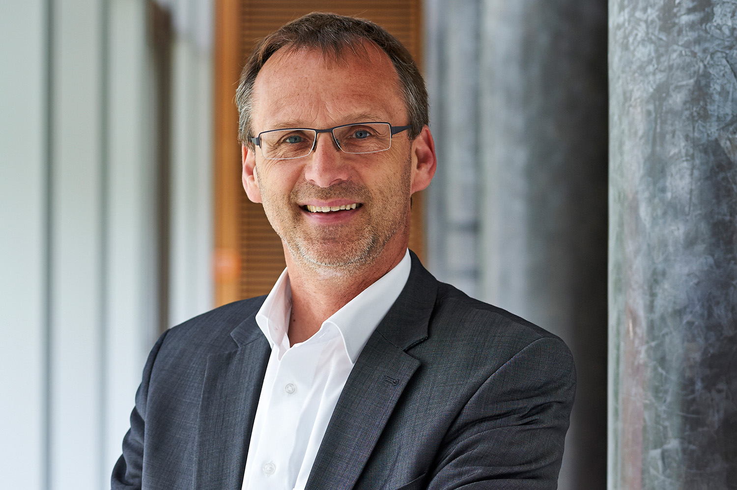 Ein Mann in Anzug und weißem Hemd steht lächelnd in einem modernen Innenraum der Hochschule Coburg. Er hat kurze Haare, trägt eine Brille und hat die Arme verschränkt. Im Hintergrund sind Säulen und verschwommene Elemente zu sehen, die an eine Büroumgebung erinnern.