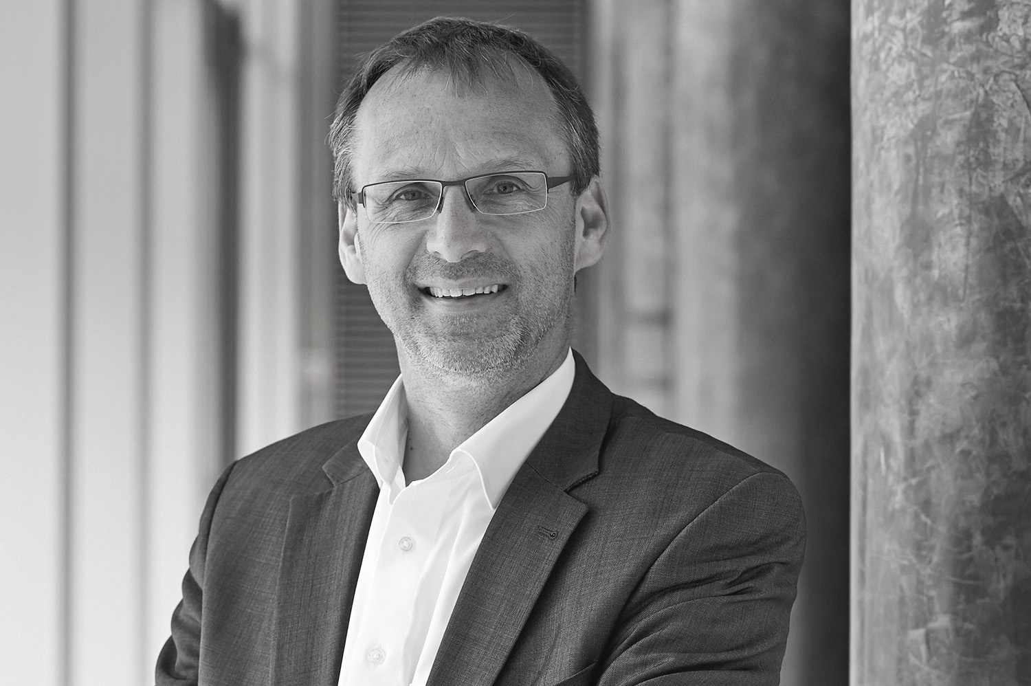 Ein Graustufenbild zeigt einen Mann im Anzug, mit Brille und strahlend weißem Hemd. Er steht selbstbewusst mit verschränkten Armen vor einer Innensäule der Hochschule Coburg und sein Lächeln strahlt Wärme und Professionalität aus.