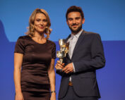 Auf der Bühne der Hochschule Coburg vor blauem Hintergrund steht eine Frau im dunklen Kleid neben einem Mann im Anzug und hält eine Trophäe in der Hand. Beide lächeln in die Kamera.