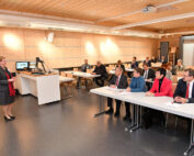 Eine Frau steht vor einem Hörsaal der Hochschule Coburg und spricht zu einer Gruppe sitzender Männer und Frauen. Der Raum ist mit Holz verkleidet und modern gestaltet. Die Teilnehmer hören aufmerksam zu, manche machen sich Notizen, während ein Projektor und eine Leinwand wichtige Informationen anzeigen.