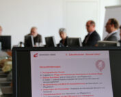 In einem Besprechungsraum der Hochschule Coburg sitzt eine Gruppe von Menschen an einem Konferenztisch und diskutiert über Themen, die auf einem Bildschirm angezeigt werden. Auf dem Bildschirm sind verschiedene Herausforderungen für Gesundheit und Pflege aufgelistet, darunter der demografische Wandel und digitale Lösungen.