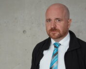 Ein kahlköpfiger Mann mit rötlichem Voll- und Schnurrbart, bekleidet mit weißem Hemd, gestreifter türkisfarbener Krawatte und schwarzem Jackett, steht vor einer grauen Betonwand. Er blickt mit neutralem Gesichtsausdruck direkt in die Kamera und verkörpert damit den professionellen Geist der Hochschule Coburg.