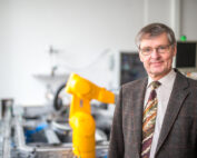 Ein Mann in Anzug und Krawatte steht selbstbewusst in einem Labor der Hochschule Coburg, im Hintergrund ein gelber Roboterarm. Sein graues Haar und seine Brille ergänzen sein leichtes Lächeln. Der Raum ist verschwommen und lenkt die Aufmerksamkeit sowohl auf ihn als auch auf die fortschrittliche Technologie.