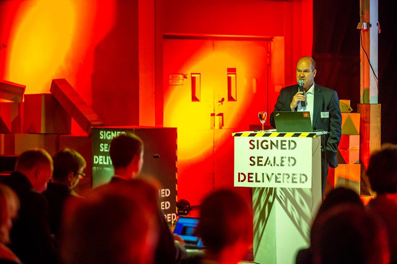 Ein Mann von der Hochschule Coburg spricht an einem Rednerpult mit der Aufschrift „Signed Sealed Delivered“ in einem schwach beleuchteten Raum mit orangefarbener und roter Beleuchtung. Ein aufmerksames Publikum hört sitzend zu, im Hintergrund sind große Kartons gestapelt.