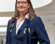 Eine Person mit langen Haaren, Brille, blauem Anzug, weißem Hemd und marineblauem Schal steht selbstbewusst im modernen Innenraum der Hochschule Coburg, beleuchtet von einem kreisförmigen Lichtelement darüber.