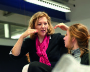 Eine Frau mit einem rosa Schal gestikuliert lebhaft, während sie mit einer anderen Frau mit zurückgebundenem Haar spricht. Wahrscheinlich diskutieren sie über ein Projekt an der Hochschule Coburg. Ihre Interaktion steht im Mittelpunkt, wobei eine verschwommene Flasche und Deckenlichter den Hintergrund in dieser lebhaften Innenumgebung bilden.