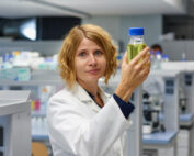 In einem modernen Labor der Hochschule Coburg hält eine Wissenschaftlerin im Laborkittel einen Behälter mit gelbgrüner Flüssigkeit hoch. Ihr kurzes blondes Haar umrahmt ihren konzentrierten Gesichtsausdruck, während sie die Substanz genau beobachtet, umgeben von hochmodernen wissenschaftlichen Geräten und Materialien.