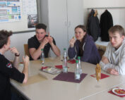 Vier junge Erwachsene sitzen an einem Tisch in der Hochschule Coburg und genießen einen gemütlichen Snack. Auf dem Tisch stehen Wasserflaschen, Brezeln und Teller mit Essen. Sie lächeln und scheinen in ein Gespräch vertieft zu sein. Im Hintergrund sind eine Garderobe und ein helles Poster zu sehen.