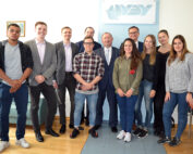 Eine Gruppe von elf Personen, sowohl Männer als auch Frauen, steht in einem Büro der Hochschule Coburg zusammen. Sie lächeln und tragen eine Mischung aus Freizeit- und Businesskleidung. Ein Logo an der Wand hinter ihnen lautet „QXY“.