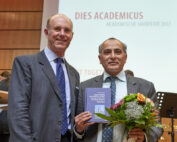 Zwei Männer in Anzügen stehen zusammen und lächeln herzlich. Einer hält ein Buch und einen Blumenstrauß in der Hand. Hinter ihnen ist auf einem Präsentationsbildschirm der Titel „Dies Academicus Akademische Jahresfeier 2017“ zu sehen, der den Geist der Hochschule Coburg feiert.