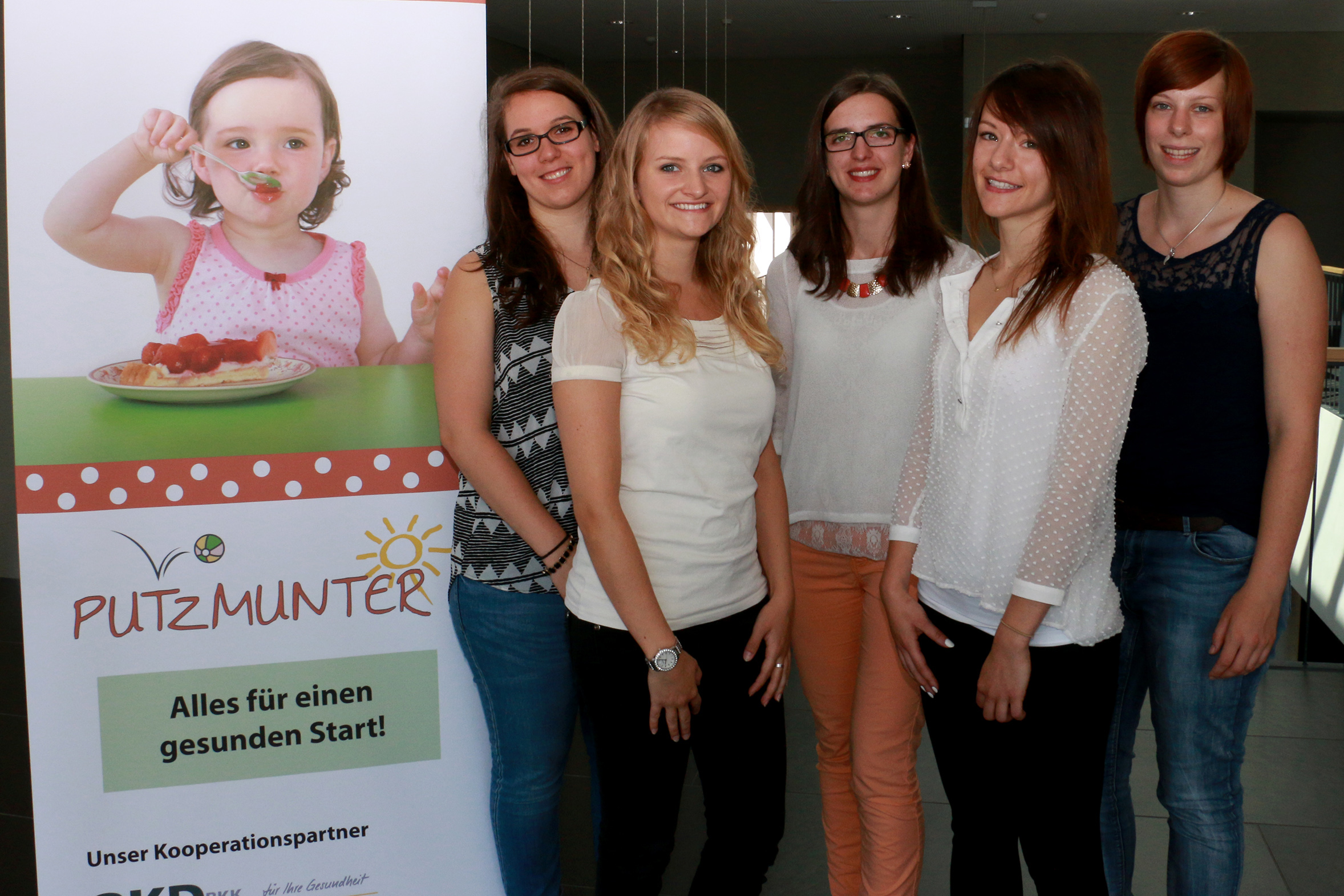 Fünf Frauen stehen drinnen neben einem Werbebanner der Hochschule Coburg, auf dem ein Kind Gemüse isst. Auf dem Banner steht „PUTZMUNTER“ und „Alles für einen gesunden Start!“. Die Frauen sind leger gekleidet und lächeln und strahlen Wärme und Positivität aus.