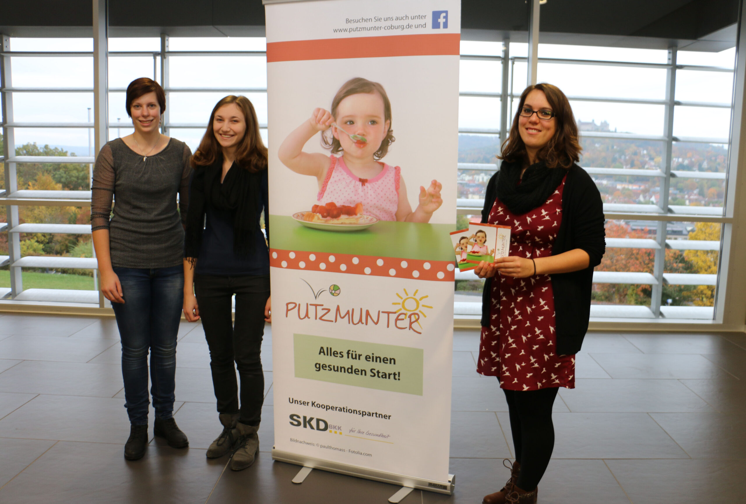 Drei Personen stehen neben einem Banner, auf dem ein Kind beim Gemüseessen zu sehen ist, mit der Aufschrift „PUTZMUNTER Alles für einen gesunden Start!“ in einem hellen, modernen Raum mit großen Fenstern an der Hochschule Coburg.