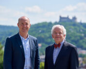 Zwei Männer in Anzügen stehen im Freien. Im Hintergrund sind ein Hügel und das historische Schloss der Hochschule Coburg zu sehen. Sie lächeln beide und der Himmel ist klar.