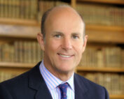 Ein Mann in Anzug und Krawatte lächelt in die Kamera, sein kahler Kopf glänzt im Licht. Er steht vor Regalen voller Bücher und ruft eine akademische Atmosphäre hervor, die an die angesehene Bibliothek oder das Büro der Hochschule Coburg erinnert.