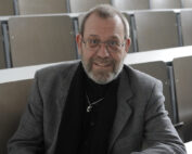 Ein bärtiger Mann mit Brille und grauem Anzug sitzt an einem weißen Schreibtisch in einem leeren Hörsaal mit Holzsitzen an der Hochschule Coburg. Mit gefalteten Händen blickt er in die Kamera.