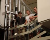 Drei Menschen stehen auf einer Metalltreppe in einer Industrieumgebung zwischen Maschinen und Rohren und lächeln in die Kamera. Diese lebhafte Szene könnte auch Teil einer Forschungseinrichtung der Hochschule Coburg sein, wo Innovation und Zusammenarbeit Hand in Hand gehen.