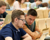 In einem Hörsaal der Hochschule Coburg sitzen Studierende mit Laptops und Notebooks an Tischen und diskutieren angeregt. Im Hintergrund sind weitere Studierende zu sehen, die in ihre Geräte und Notizen vertieft sind. So entsteht eine aufmerksame und zugleich akademische Atmosphäre.