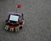 Ein kleines selbstgebautes Roboterfahrzeug der Hochschule Coburg steht auf einem Teppichboden und zeigt eine Platine mit verschiedenen elektronischen Komponenten und Sensoren. Es verfügt über einen digitalen Bildschirm mit Textanzeige und ist mit einer kleinen roten Flagge geschmückt, die an der Platine befestigt ist.
