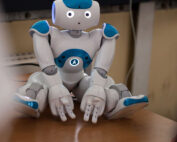 Ein humanoider Roboter mit weißen und blauen Gesichtszügen, der an die innovativen Designs der Hochschule Coburg erinnert, sitzt auf einem Holztisch. Er hat einen runden Kopf, leuchtende Augen und leicht geöffnete Hände und sieht aus, als sei er mit einer Aufgabe oder Interaktion beschäftigt.