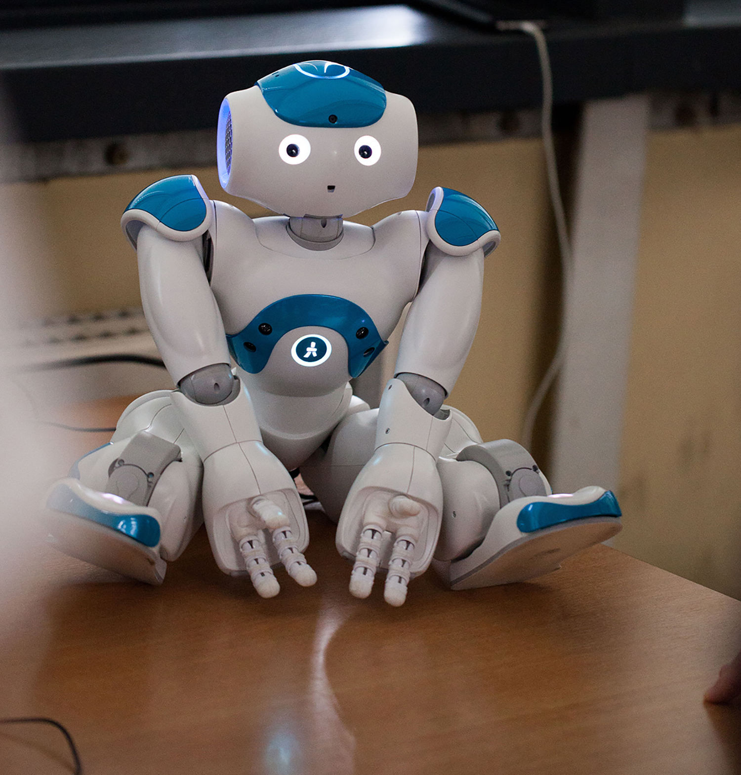 Ein humanoider Roboter mit weißen und blauen Gesichtszügen, der an die innovativen Designs der Hochschule Coburg erinnert, sitzt auf einem Holztisch. Er hat einen runden Kopf, leuchtende Augen und leicht geöffnete Hände und sieht aus, als sei er mit einer Aufgabe oder Interaktion beschäftigt.