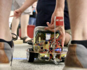 Eine Person kniet, um einen kleinen Roboter auf dem Boden einzustellen, umgeben von anderen in Freizeitkleidung an der Hochschule Coburg. Der Roboter mit sichtbaren Kabeln und Komponenten steht neben einer blauen Linie und auf einer kleinen Flagge steht „AC-motiv“.