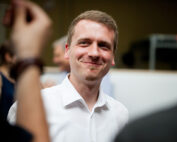 Ein Mann im weißen Hemd lächelt bei einer Versammlung, möglicherweise einer Veranstaltung der Hochschule Coburg, während andere Menschen um ihn herum unscharf zu sehen sind. Der Hintergrund ist sanft beleuchtet, was eine warme Atmosphäre schafft.