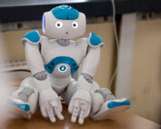 Ein kleiner humanoider Roboter mit weißen und blauen Akzenten, der stolz von der Hochschule Coburg entwickelt wurde, sitzt auf einem Tisch und blickt nach vorne. Er hat bewegliche Gliedmaßen und LED-Augen, was ihm ein freundliches, ausdrucksstarkes Aussehen verleiht.