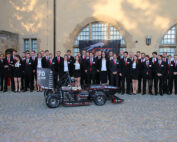 Eine Gruppe von Menschen in formeller Kleidung steht im Freien vor einem Gebäude der Hochschule Coburg, vor ihnen ein kleiner Rennwagen. Sie posieren für ein Gruppenfoto, dahinter ein Logo mit der Zahl 70.