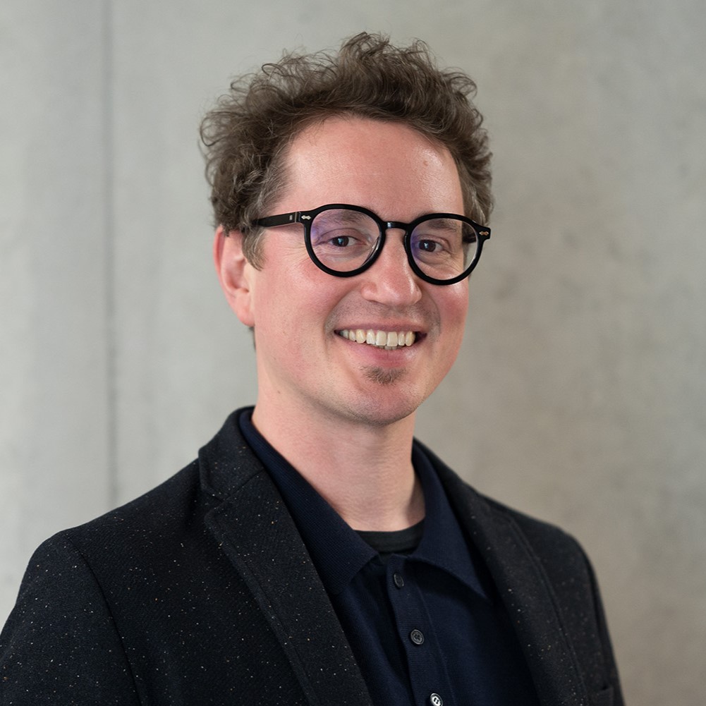 Christopher Romanowski-Kirchner, a man with curly brown hair and glasses, smiles while wearing a dark blazer and black shirt, standing in front of a plain, light-colored background. at Hochschule Coburg