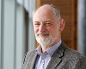 Ein bärtiger Mann mit grauem Bart und Haar lächelt und trägt einen grauen Blazer über einem gestreiften Hemd. Er steht im Innenbereich der Hochschule Coburg, eingerahmt von großen Fenstern und einer Holzverkleidung.