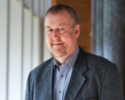 Ein Mann mit kurzen, hellen Haaren und Bart lächelt, während er einen grauen Blazer und ein graues Hemd trägt. Er steht vor einem verschwommenen Hintergrund mit großen Fenstern zu seiner Linken an der Hochschule Coburg.